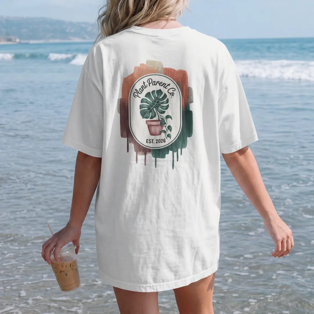 Woman on the beach in a White tee, back view: a neat oval crest with a monstera leaf and a pothos vine framing a small pot, "PLANT PARENT CO." in loose brush script above and "EST. 2026" below, painted in loose watercolor and ink with soft deep…