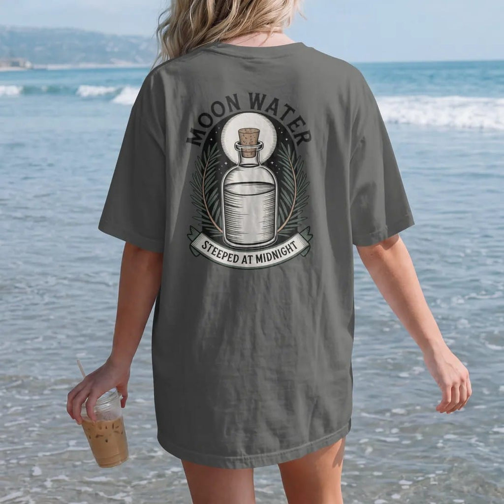 Woman on the beach in a Pepper tee, back view: back print: A corked glass bottle of water under a full moon, rosemary sprigs around it, arched header "MOON WATER" above, small banner "STEEPED AT MIDNIGHT" below, detailed vintage etching, fine…