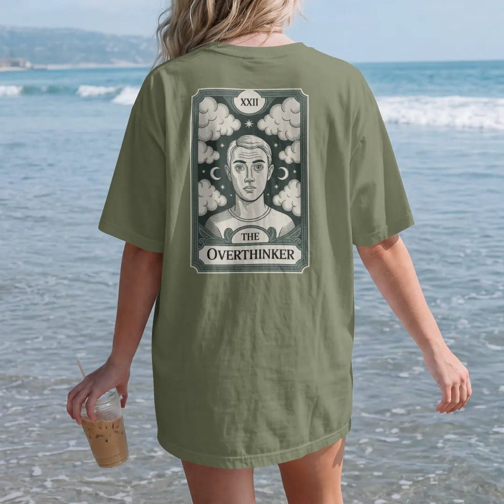 Woman on the beach in a Moss tee, back view: back print: A tarot card frame with a wide-eyed figure surrounded by swirling thought clouds and tiny moons, Roman numeral "XXII" at the top, nameplate "THE OVERTHINKER" at the bottom, antique…