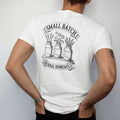 Man wearing a White tee, seen from the back: back print: three apothecary jars with blank labels holding lavender, chamomile and mugwort on a wooden shelf, curved header "SMALL BATCH" above, ribbon "HERBAL REMEDIES" below, detailed vintage etching,…