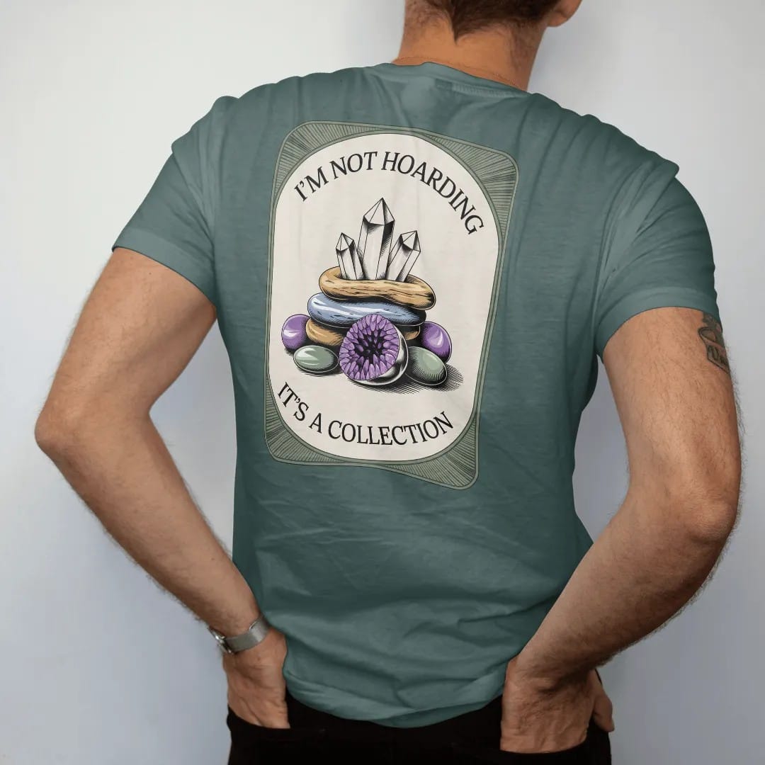 Man wearing a Blue Spruce tee, seen from the back: back print: A heap of quartz points, amethyst geodes and tumbled stones, stacked text "I'M NOT HOARDING" above and "IT'S A COLLECTION" below the pile, antique apothecary-label engraving, fine…