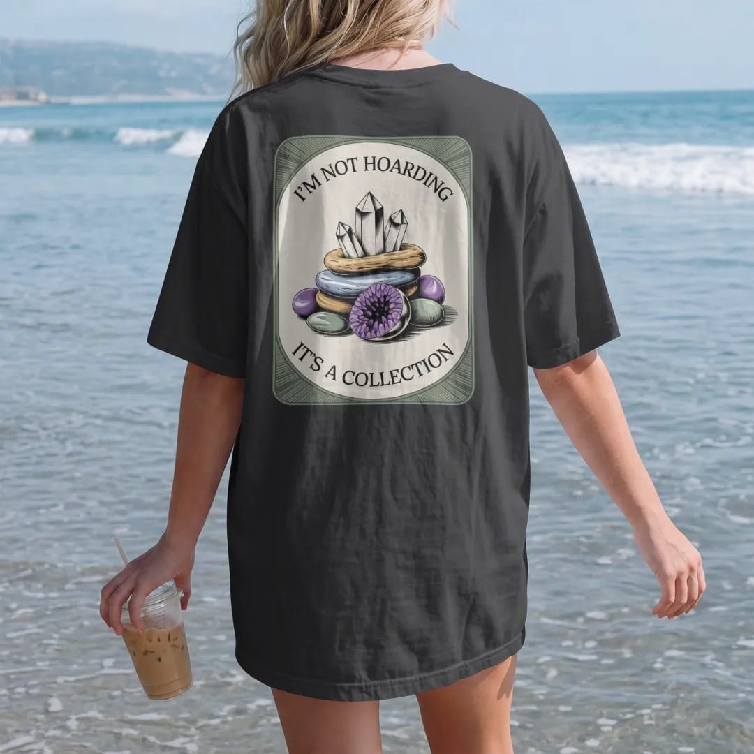 Woman on the beach in a Black tee, back view: back print: A heap of quartz points, amethyst geodes and tumbled stones, stacked text "I'M NOT HOARDING" above and "IT'S A COLLECTION" below the pile, antique apothecary-label engraving, fine…