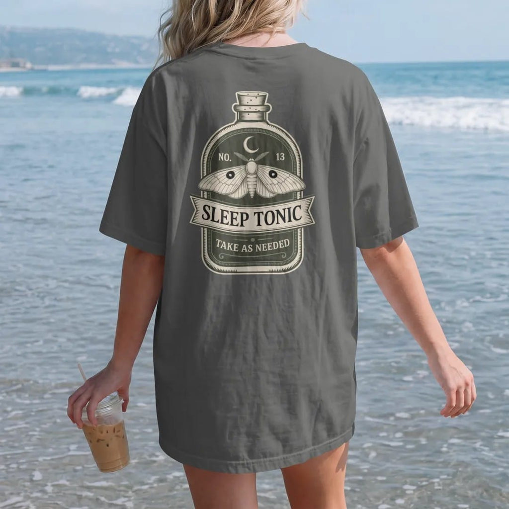 Woman on the beach in a Pepper tee, back view: back print: A vintage potion bottle label with a sleeping moth and a crescent moon, label layout with "NO. 13" on top, "SLEEP TONIC" in the center banner and "TAKE AS NEEDED" below, detailed vintage…