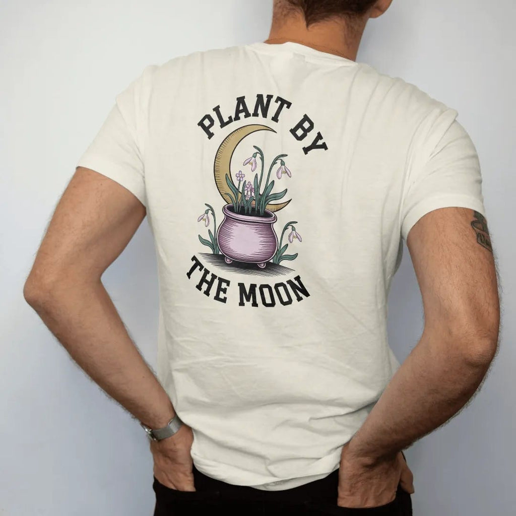 Man wearing a Ivory tee, seen from the back: back print: seedlings sprouting from a little cauldron-shaped pot under a waxing crescent, with snowdrops and violets, arched "PLANT BY" over the top and "THE MOON" curved beneath, detailed vintage…