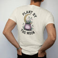 Man wearing a Ivory tee, seen from the back: back print: seedlings sprouting from a little cauldron-shaped pot under a waxing crescent, with snowdrops and violets, arched "PLANT BY" over the top and "THE MOON" curved beneath, detailed vintage…