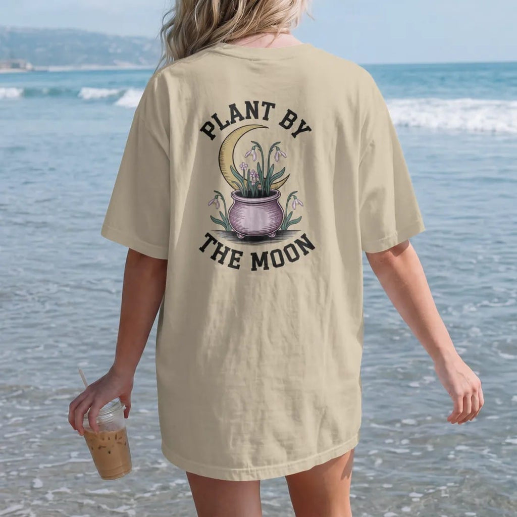 Woman on the beach in a Sandstone tee, back view: back print: seedlings sprouting from a little cauldron-shaped pot under a waxing crescent, with snowdrops and violets, arched "PLANT BY" over the top and "THE MOON" curved beneath, detailed vintage…