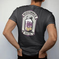 Man wearing a Black tee, seen from the back: back print: A big glass jar of sun tea with floating chamomile and lemon slices, radiating sun rays, arched "MIDSUMMER" above and small banner "SUN TEA" below, antique apothecary-label engraving, fine…