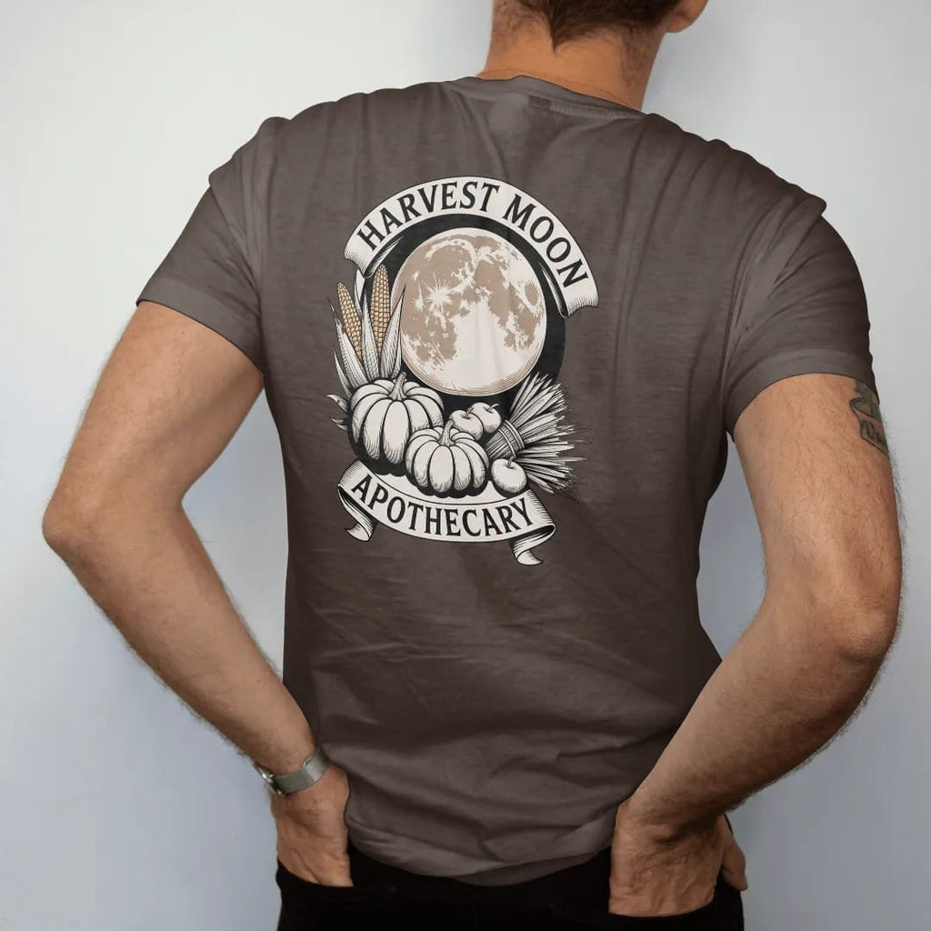 Man wearing a Espresso tee, seen from the back: back print: pumpkins, dried corn, bundled sage and apples beneath a huge harvest moon, curved "HARVEST MOON" above and "APOTHECARY" below in a ribbon, detailed vintage etching, fine crosshatching and…