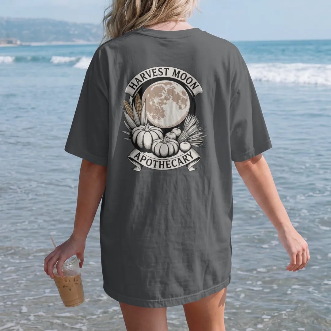 Woman on the beach in a Graphite tee, back view: back print: pumpkins, dried corn, bundled sage and apples beneath a huge harvest moon, curved "HARVEST MOON" above and "APOTHECARY" below in a ribbon, detailed vintage etching, fine crosshatching and…