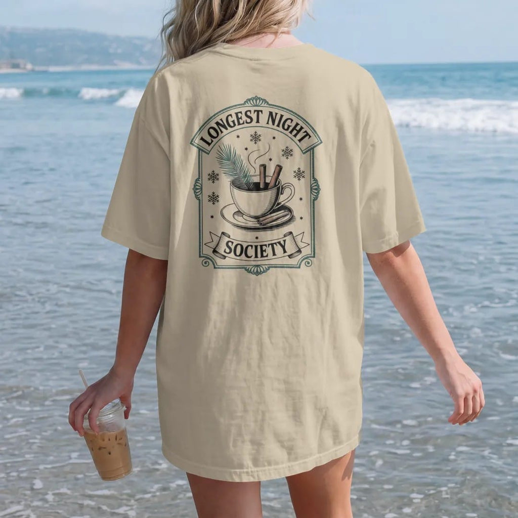 Woman on the beach in a Sandstone tee, back view: back print: A steaming teacup with a pine sprig and cinnamon sticks, surrounded by snowflakes and tiny stars, arched "LONGEST NIGHT" over the cup and "SOCIETY" in a small banner below, antique…