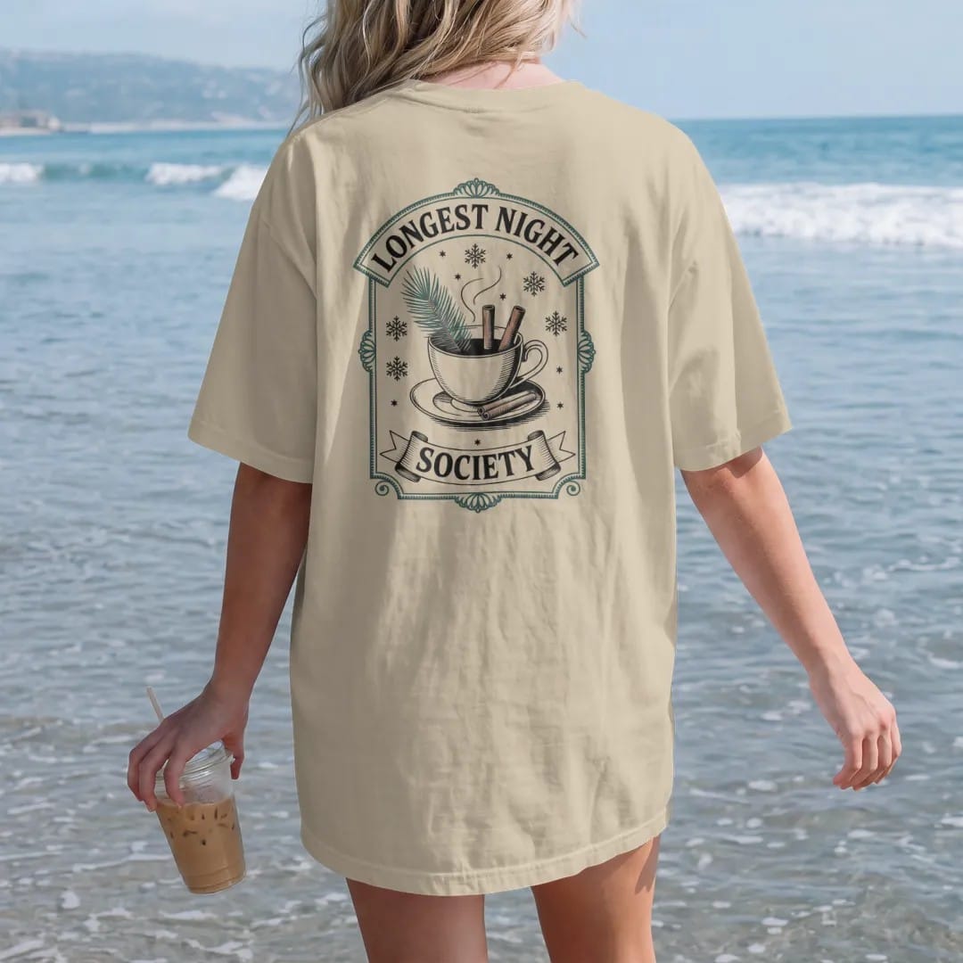 Woman on the beach in a Sandstone tee, back view: back print: A steaming teacup with a pine sprig and cinnamon sticks, surrounded by snowflakes and tiny stars, arched "LONGEST NIGHT" over the cup and "SOCIETY" in a small banner below, antique…