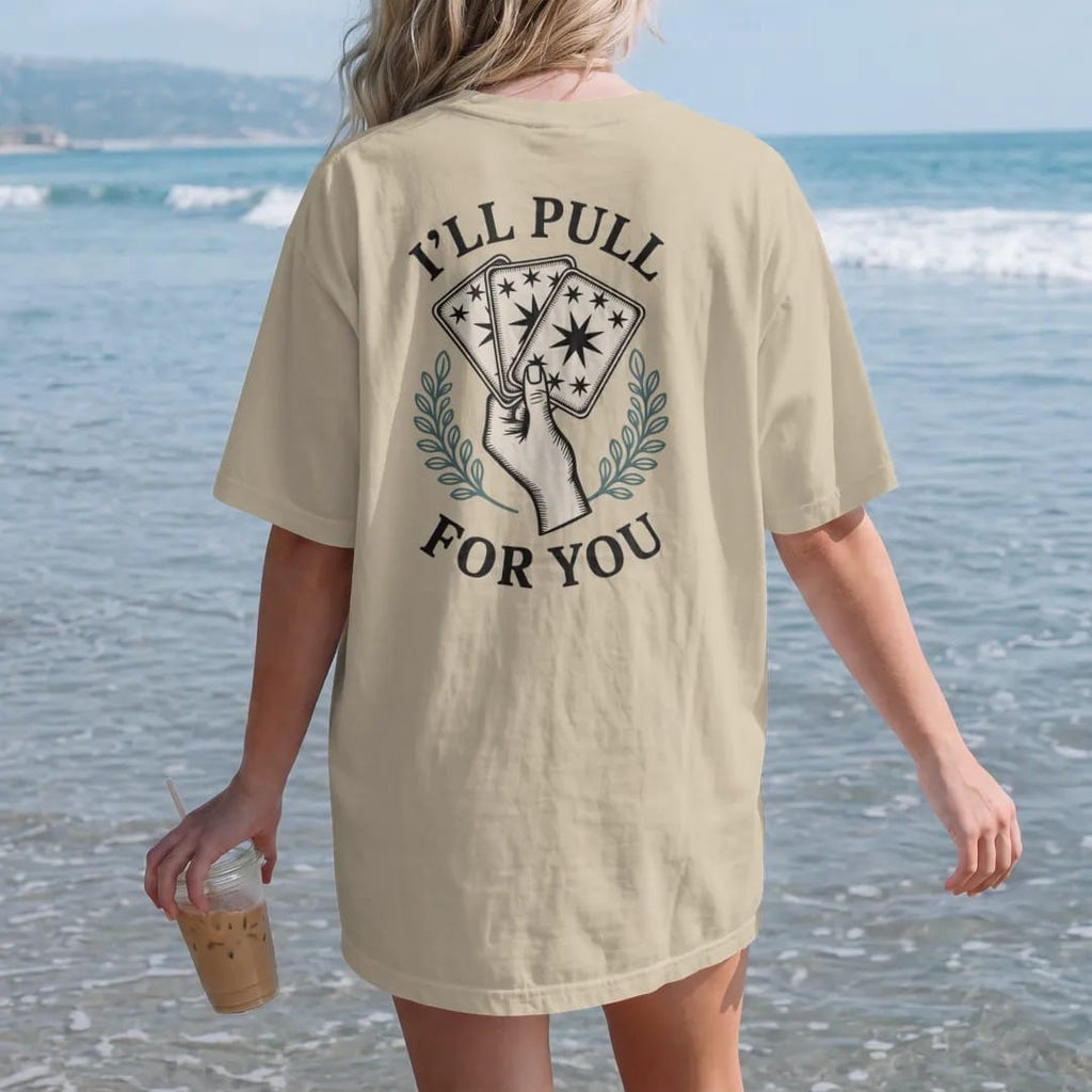 Woman on the beach in a Sandstone tee, back view: back print: A hand fanning out three face-down tarot cards with star-patterned backs, stacked text "I'LL PULL", "A CARD" and "FOR YOU" arranged around the fan, detailed vintage etching, fine…