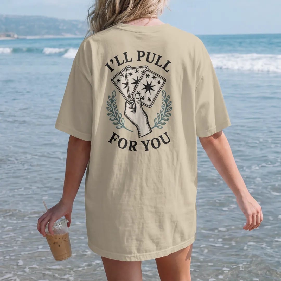 Woman on the beach in a Sandstone tee, back view: back print: A hand fanning out three face-down tarot cards with star-patterned backs, stacked text "I'LL PULL", "A CARD" and "FOR YOU" arranged around the fan, detailed vintage etching, fine…