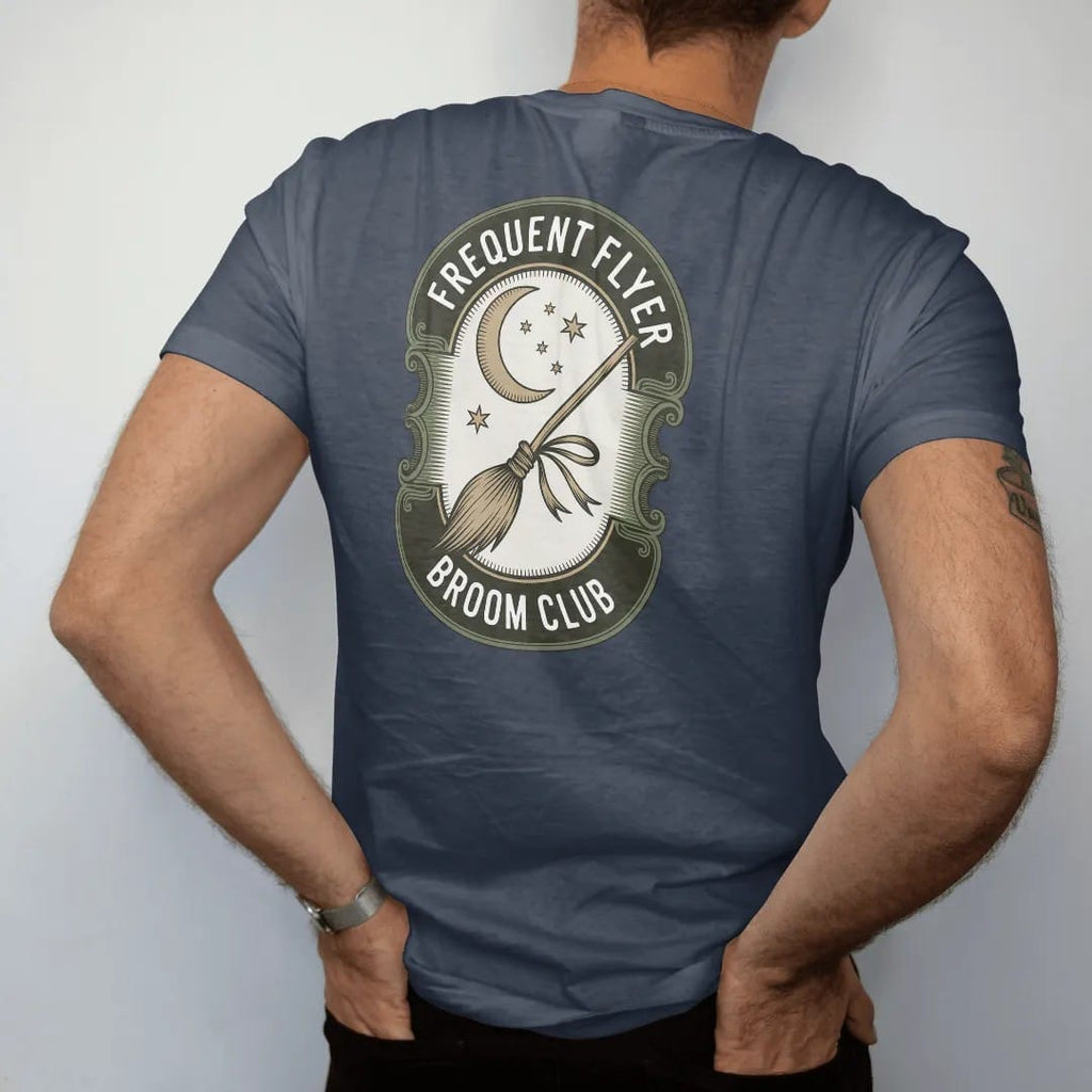 Man wearing a Midnight tee, seen from the back: back print: A vintage broomstick with a ribbon tied on, flying past a crescent moon with trailing stars, round badge with "FREQUENT FLYER" arched above and "BROOM CLUB" below, antique apothecary-label…