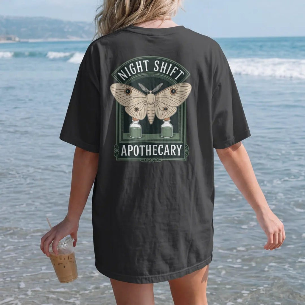 Woman on the beach in a Black tee, back view: back print: A large luna moth with wings spread over two small potion bottles, arched "NIGHT SHIFT" above and "APOTHECARY" below, antique apothecary-label engraving, fine crosshatching, ornate frame,…