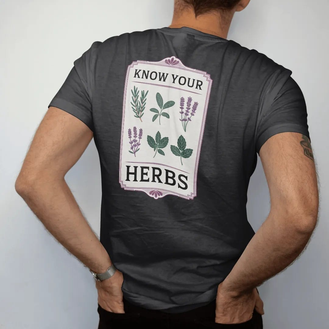 Man wearing a Black tee, seen from the back: back print: A vintage botanical chart of six sprigs in a neat grid: rosemary, sage, thyme, lavender, basil and mint, small header "KNOW YOUR" with "HERBS" in large letters, antique apothecary-label…
