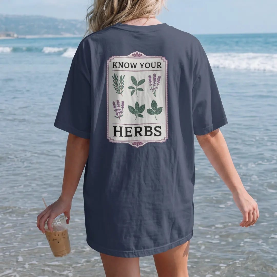 Woman on the beach in a Navy tee, back view: back print: A vintage botanical chart of six sprigs in a neat grid: rosemary, sage, thyme, lavender, basil and mint, small header "KNOW YOUR" with "HERBS" in large letters, antique apothecary-label…
