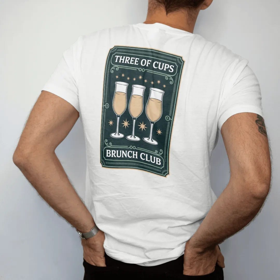 Man wearing a White tee, seen from the back: back print: A tarot card frame showing three raised mimosa glasses clinking with tiny stars, "THREE OF CUPS" at the card top, "BRUNCH CLUB" at the bottom, antique apothecary-label engraving, fine…