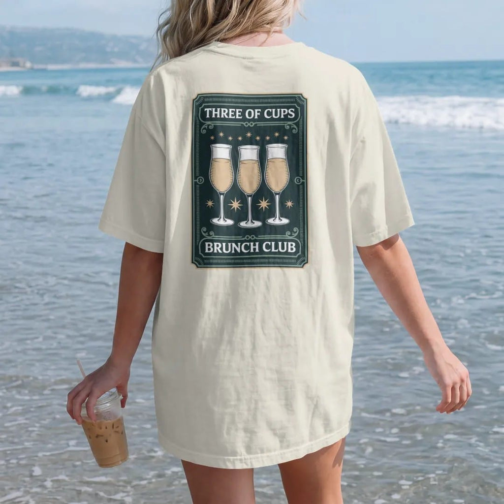 Woman on the beach in a Ivory tee, back view: back print: A tarot card frame showing three raised mimosa glasses clinking with tiny stars, "THREE OF CUPS" at the card top, "BRUNCH CLUB" at the bottom, antique apothecary-label engraving, fine…