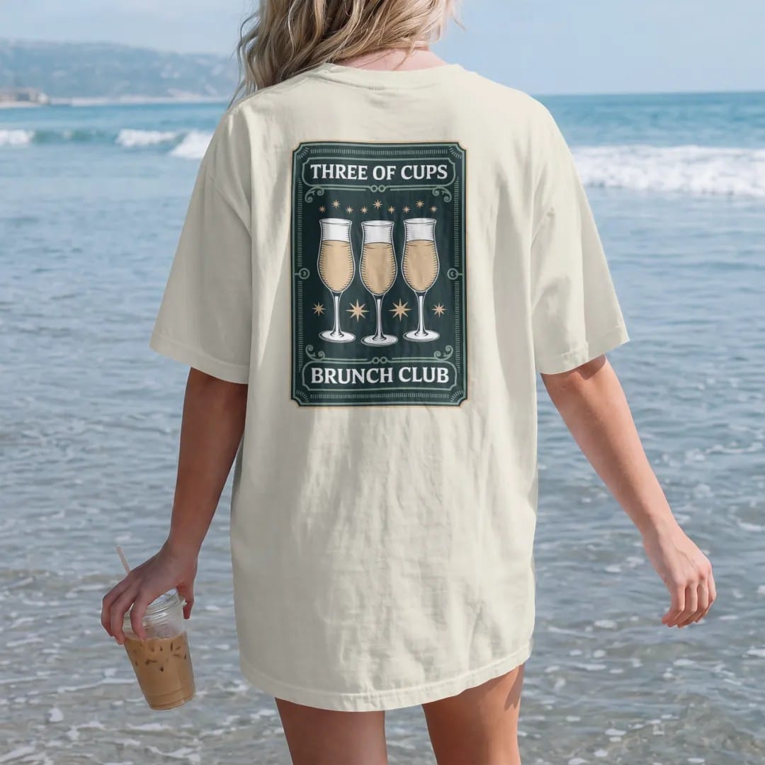Woman on the beach in a Ivory tee, back view: back print: A tarot card frame showing three raised mimosa glasses clinking with tiny stars, "THREE OF CUPS" at the card top, "BRUNCH CLUB" at the bottom, antique apothecary-label engraving, fine…