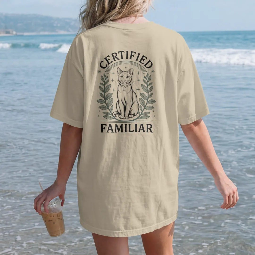 Woman on the beach in a Sandstone tee, back view: back print: A sleek cat drawn in light linework sitting upright with a crescent moon collar charm, framed by herbs and stars, arched "CERTIFIED" above and "FAMILIAR" below, detailed vintage etching,…