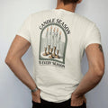 Man wearing a Ivory tee, seen from the back: back print: A cluster of tall dripping taper candles in mismatched brass holders with pooled wax, arched "CANDLE SEASON" above and "IS EVERY SEASON" below, antique apothecary-label engraving, fine…