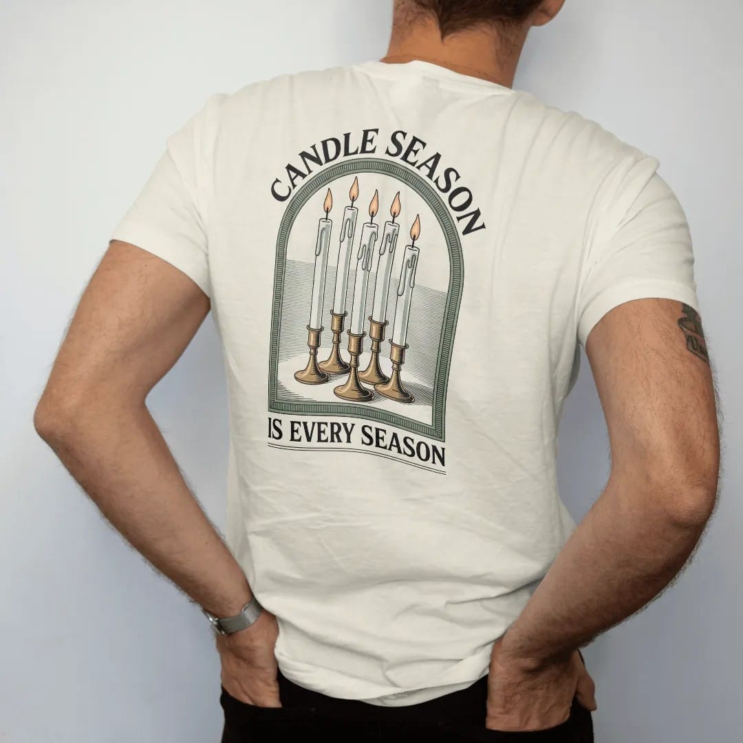 Man wearing a Ivory tee, seen from the back: back print: A cluster of tall dripping taper candles in mismatched brass holders with pooled wax, arched "CANDLE SEASON" above and "IS EVERY SEASON" below, antique apothecary-label engraving, fine…