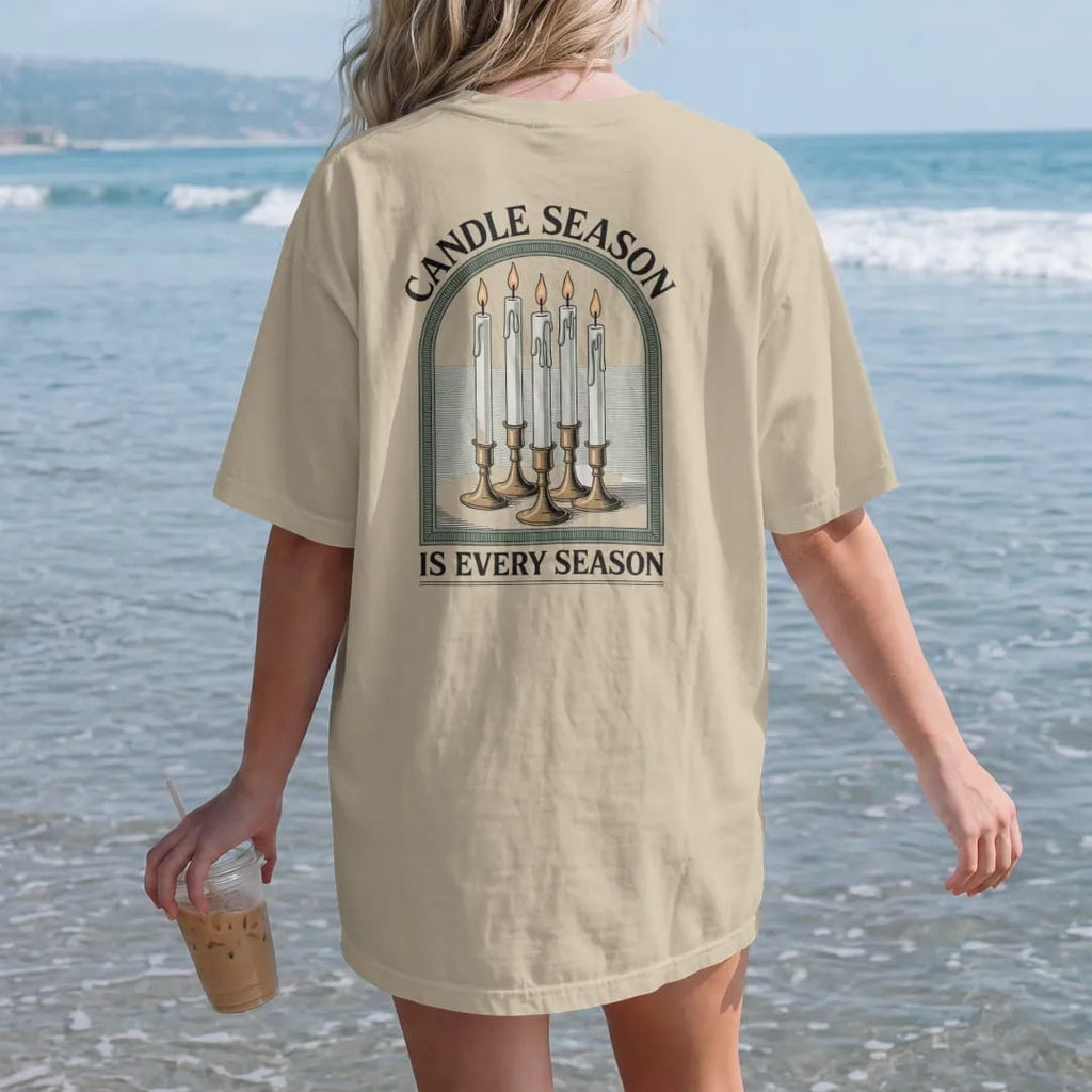 Woman on the beach in a Sandstone tee, back view: back print: A cluster of tall dripping taper candles in mismatched brass holders with pooled wax, arched "CANDLE SEASON" above and "IS EVERY SEASON" below, antique apothecary-label engraving, fine…