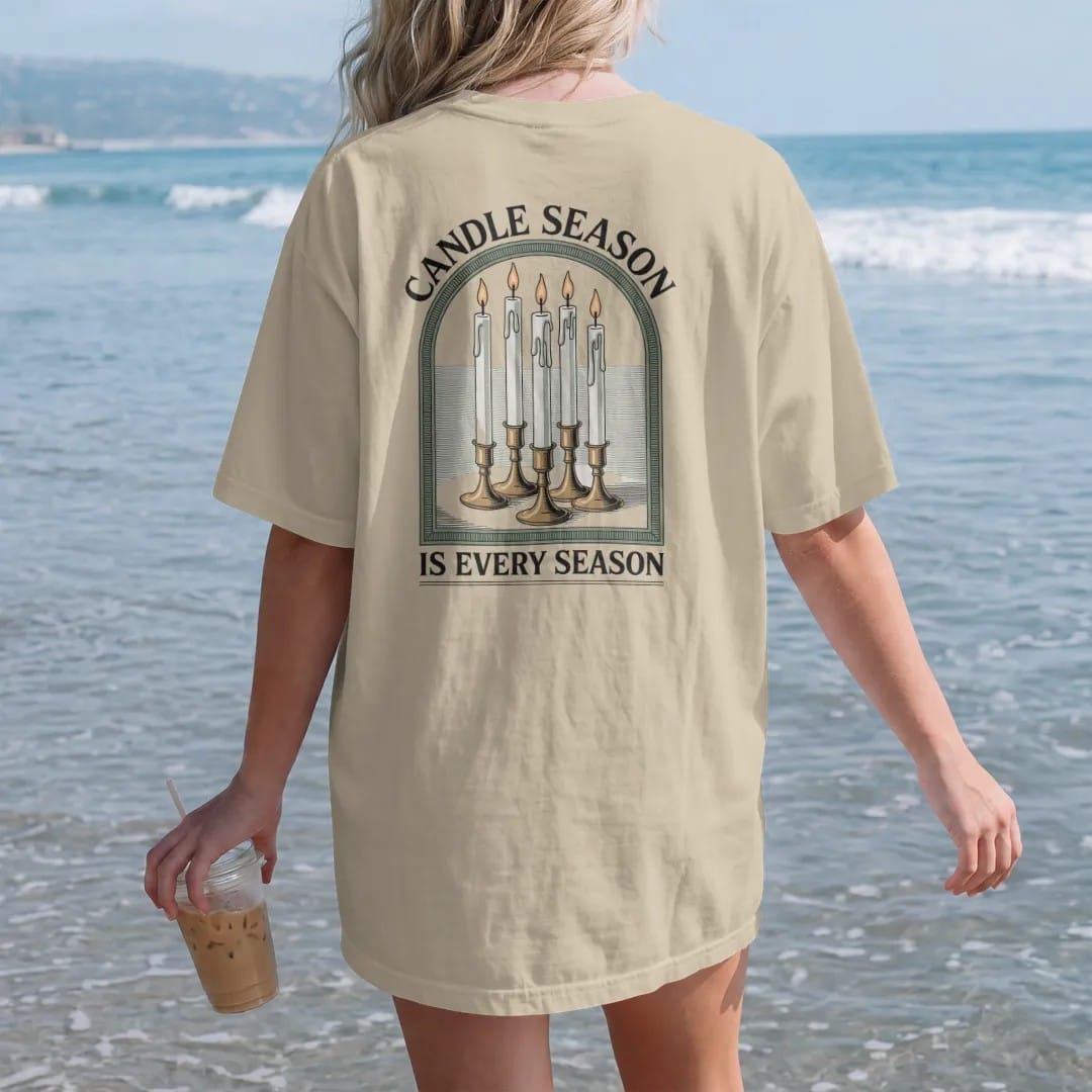 Woman on the beach in a Sandstone tee, back view: back print: A cluster of tall dripping taper candles in mismatched brass holders with pooled wax, arched "CANDLE SEASON" above and "IS EVERY SEASON" below, antique apothecary-label engraving, fine…