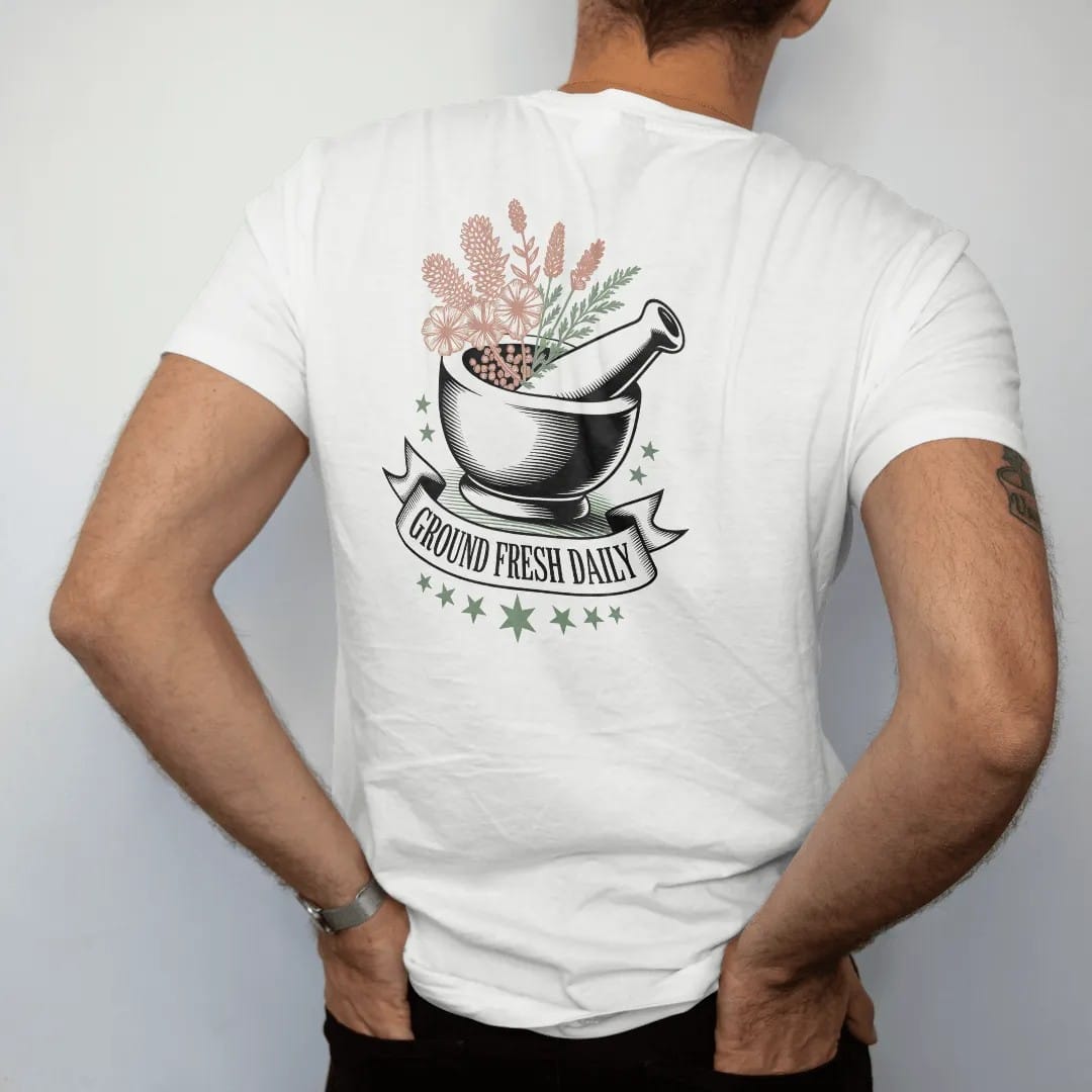 Man wearing a White tee, seen from the back: back print: A stone mortar and pestle overflowing with dried flowers, peppercorns and herbs, banner "GROUND FRESH DAILY" across the bottom with small stars around, detailed vintage etching, fine…