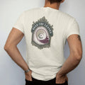 Man wearing a Ivory tee, seen from the back: back print: A porcelain teacup seen from above, tea leaves in the bottom forming a tiny crescent and star, "READ THE LEAVES" arched around the cup, antique apothecary-label engraving, fine crosshatching,…