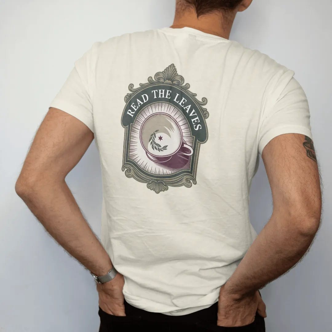 Man wearing a Ivory tee, seen from the back: back print: A porcelain teacup seen from above, tea leaves in the bottom forming a tiny crescent and star, "READ THE LEAVES" arched around the cup, antique apothecary-label engraving, fine crosshatching,…
