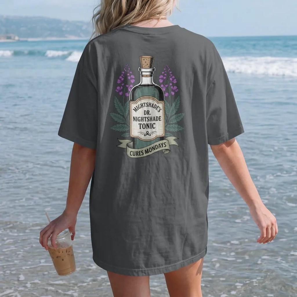 Woman on the beach in a Graphite tee, back view: back print: A vintage medicine-show bottle with a tall label, flanked by nightshade flowers, "DR. NIGHTSHADE'S" arched on top, "MIDNIGHT TONIC" on the label, "CURES MONDAYS" in a ribbon, detailed…