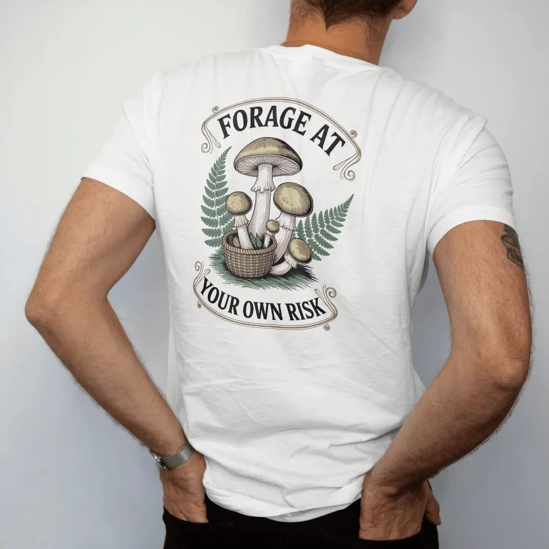 Man wearing a White tee, seen from the back: back print: A cluster of toadstools and chanterelles with a small woven basket and ferns, stacked "FORAGE AT" above and "YOUR OWN RISK" below, antique apothecary-label engraving, fine crosshatching,…