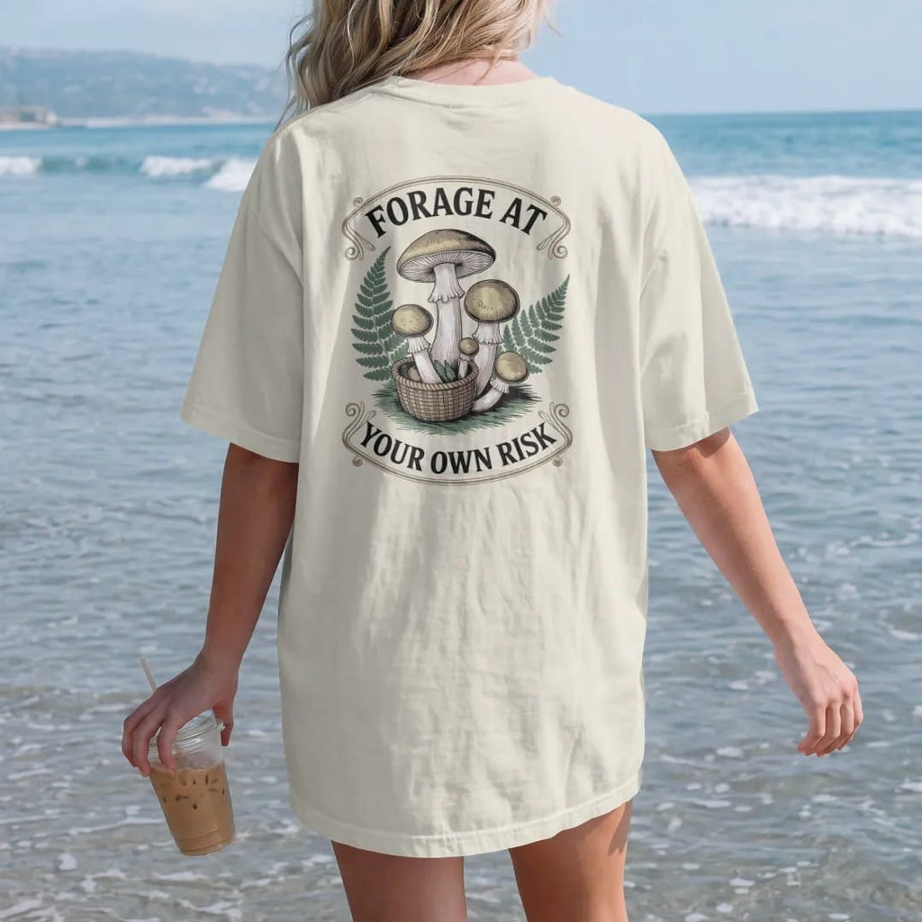Woman on the beach in a Ivory tee, back view: back print: A cluster of toadstools and chanterelles with a small woven basket and ferns, stacked "FORAGE AT" above and "YOUR OWN RISK" below, antique apothecary-label engraving, fine crosshatching,…
