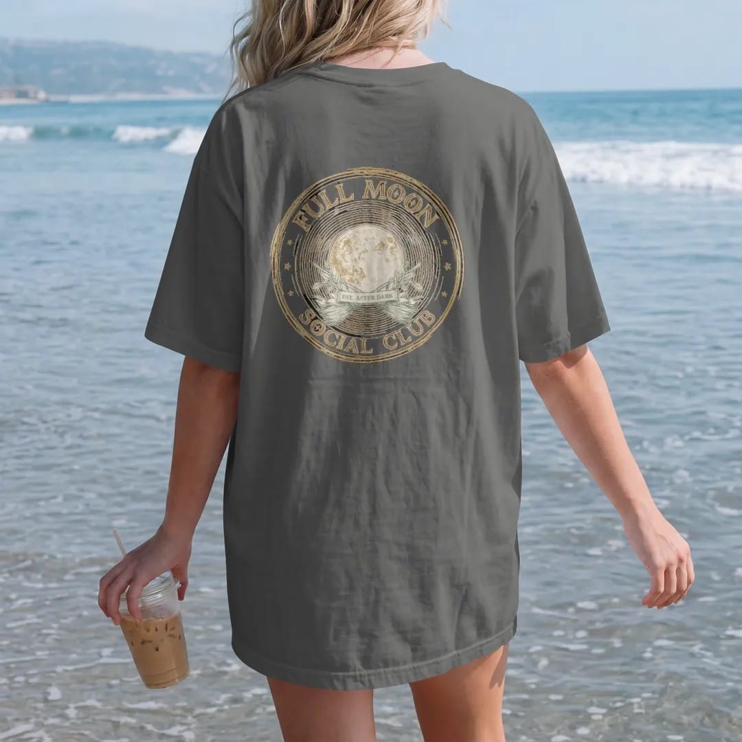 Woman on the beach in a Pepper tee, back view: back print: A club emblem with a full moon, crossed brooms and sage leaves, round badge, "FULL MOON" arched top, "SOCIAL CLUB" arched bottom, "EST. AFTER DARK" in a center ribbon, detailed vintage…