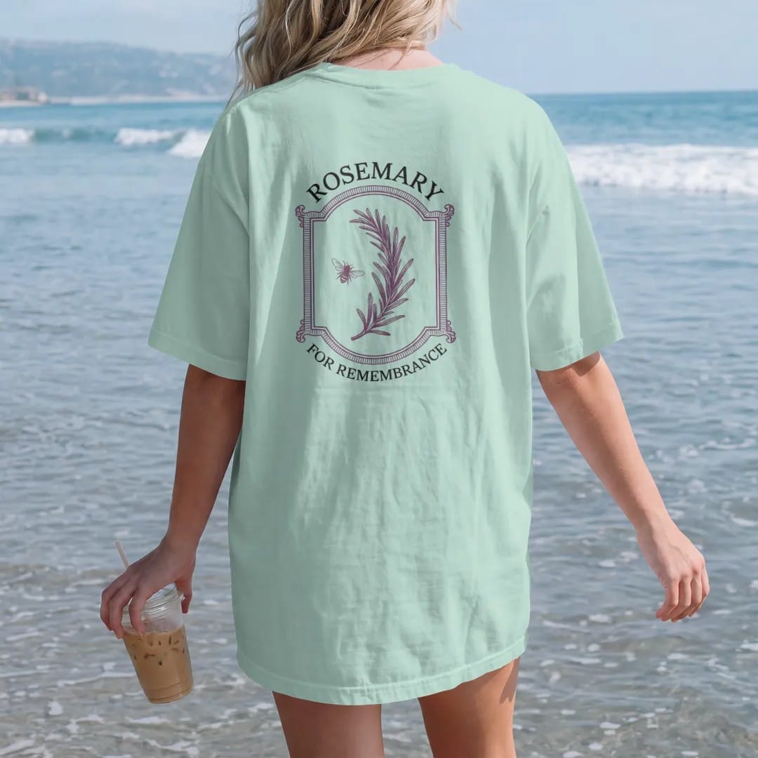 Woman on the beach in a Chalky Mint tee, back view: back print: A single long sprig of rosemary in fine engraving with a tiny bee, "ROSEMARY" above and "FOR REMEMBRANCE" in small caps below, antique apothecary-label engraving, fine crosshatching,…