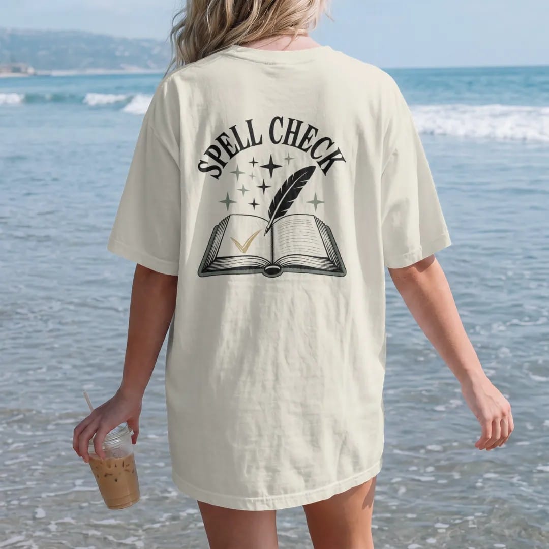 Woman on the beach in a Ivory tee, back view: back print: an open spellbook with a quill and a glowing checkmark on the page, stars rising from it, arched "SPELL CHECK" above the book, detailed vintage etching, fine crosshatching and stippling,…