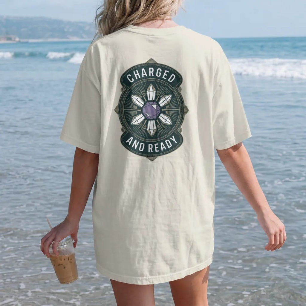 Woman on the beach in a Ivory tee, back view: back print: A geometric crystal grid of six quartz points around a central amethyst joined by fine lines, small "CHARGED" above and "AND READY" below, antique apothecary-label engraving, fine…