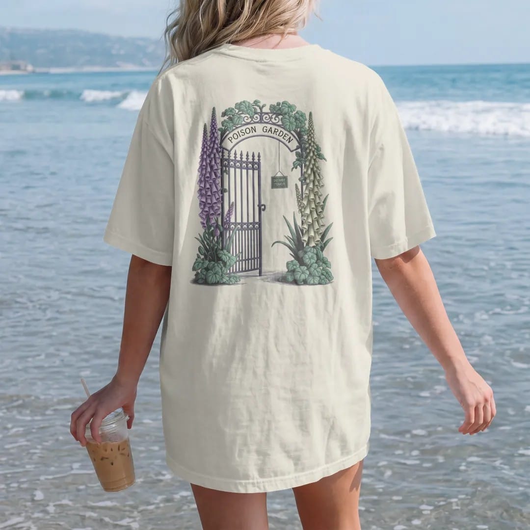 Woman on the beach in a Ivory tee, back view: back print: an iron garden gate wrapped in foxglove, belladonna and hemlock blossoms, "POISON GARDEN" arched over the gate and "DO NOT TOUCH" on a tiny hanging sign, detailed vintage etching, fine…