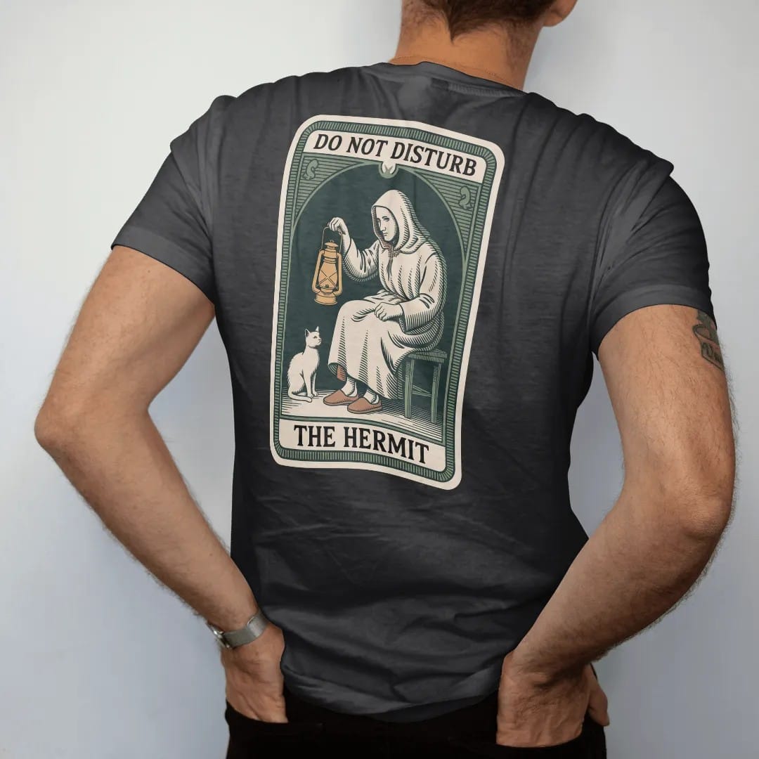 Man wearing a Black tee, seen from the back: back print: A tarot card frame with a cloaked figure holding a lantern, at home in slippers beside a cat, "DO NOT DISTURB" at the card top, "THE HERMIT" at the bottom, antique apothecary-label engraving,…