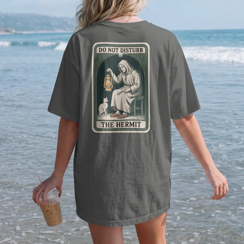 Woman on the beach in a Pepper tee, back view: back print: A tarot card frame with a cloaked figure holding a lantern, at home in slippers beside a cat, "DO NOT DISTURB" at the card top, "THE HERMIT" at the bottom, antique apothecary-label…