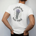 Man wearing a White tee, seen from the back: back print: A bundle of dried sage and lavender tied with string, its smoke curling into swirls and small stars, arched "CLEARING" above and "BAD VIBES" below, detailed vintage etching, fine…