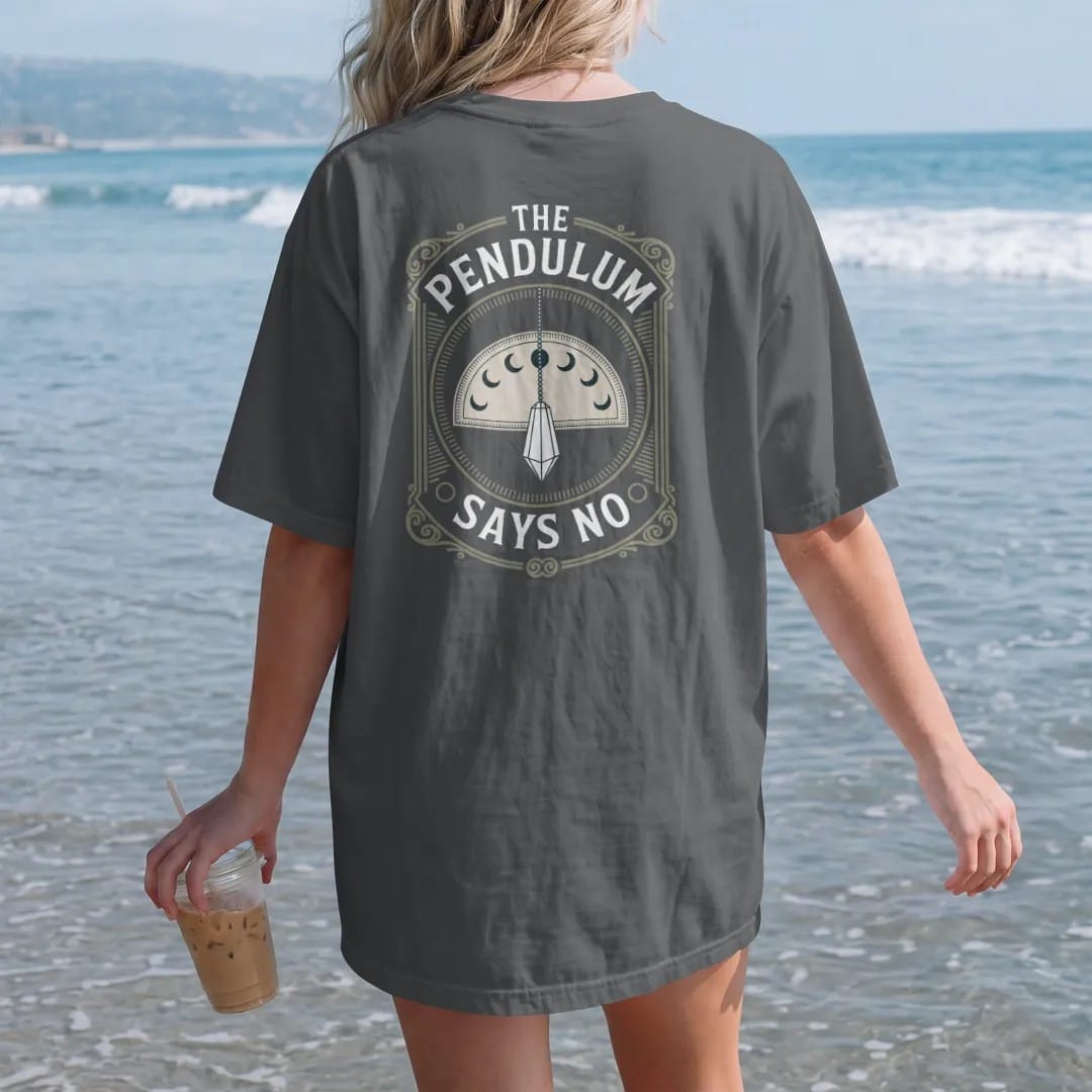 Woman on the beach in a Graphite tee, back view: back print: A crystal pendulum on a fine chain swinging over a blank half-circle board with moon symbols, stacked "THE PENDULUM" above and "SAYS NO" below, antique apothecary-label engraving, fine…