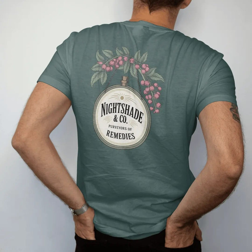 Man wearing a Blue Spruce tee, seen from the back: back print: A vintage hanging apothecary shop sign with a dangling bottle, framed by nightshade flowers and berries, hanging sign with "NIGHTSHADE & CO." large and "PURVEYORS OF REMEDIES" below,…