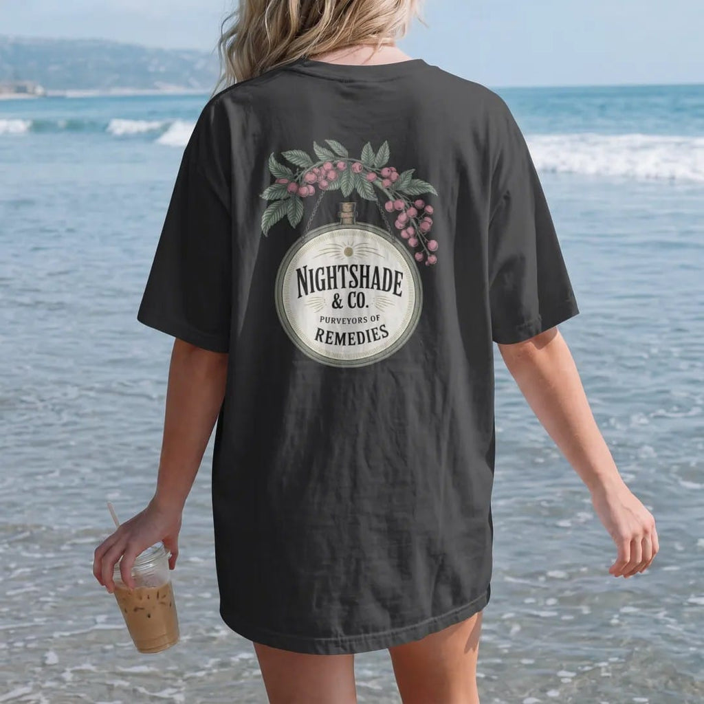 Woman on the beach in a Black tee, back view: back print: A vintage hanging apothecary shop sign with a dangling bottle, framed by nightshade flowers and berries, hanging sign with "NIGHTSHADE & CO." large and "PURVEYORS OF REMEDIES" below,…