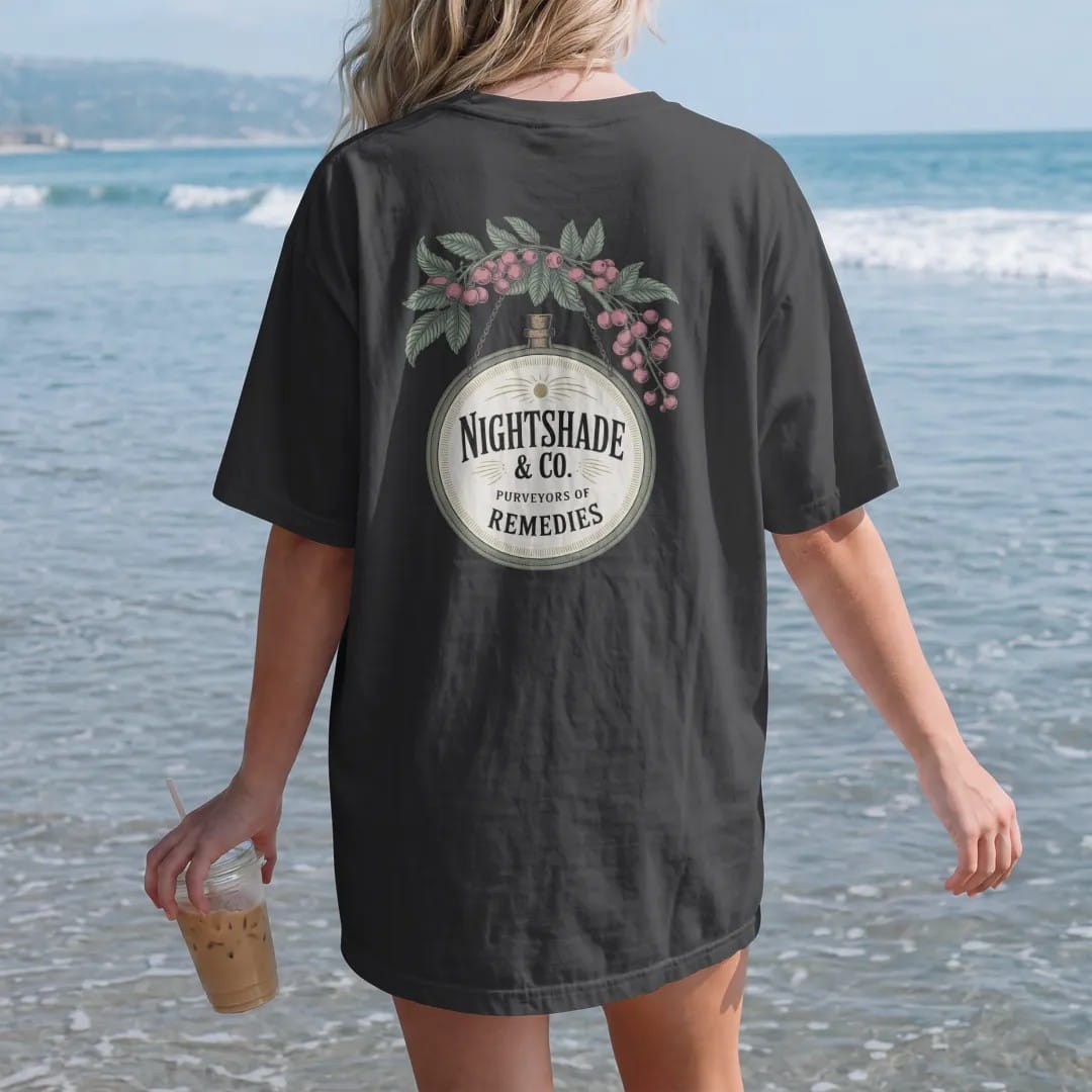 Woman on the beach in a Black tee, back view: back print: A vintage hanging apothecary shop sign with a dangling bottle, framed by nightshade flowers and berries, hanging sign with "NIGHTSHADE & CO." large and "PURVEYORS OF REMEDIES" below,…