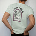Man wearing a Bay tee, seen from the back: back print: A sleeping crescent moon face wrapped in a garland of wildflowers and stars, soft arch "MOON CHILD" above, antique apothecary-label engraving, fine crosshatching, ornate frame, fine copperplate…