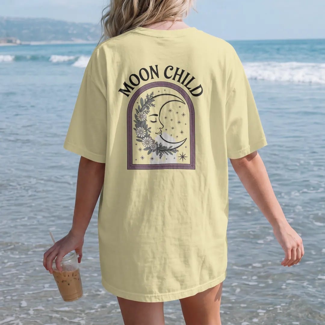 Woman on the beach in a Butter tee, back view: back print: A sleeping crescent moon face wrapped in a garland of wildflowers and stars, soft arch "MOON CHILD" above, antique apothecary-label engraving, fine crosshatching, ornate frame, fine…