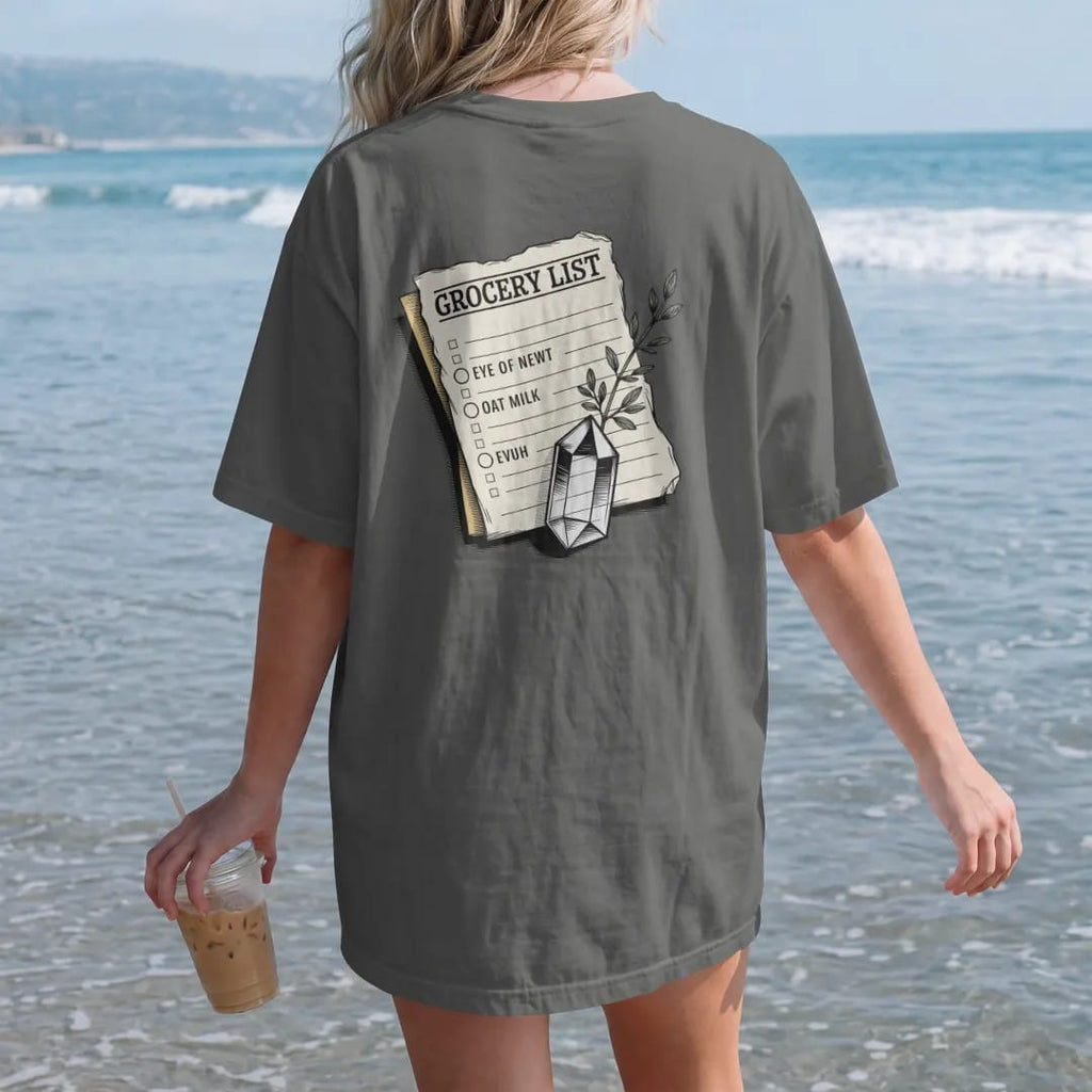 Woman on the beach in a Pepper tee, back view: back print: A torn vintage paper note held down by a small quartz crystal, with a sprig of thyme, list layout with header "GROCERY LIST" and the lines "EYE OF NEWT" and "OAT MILK", detailed vintage…